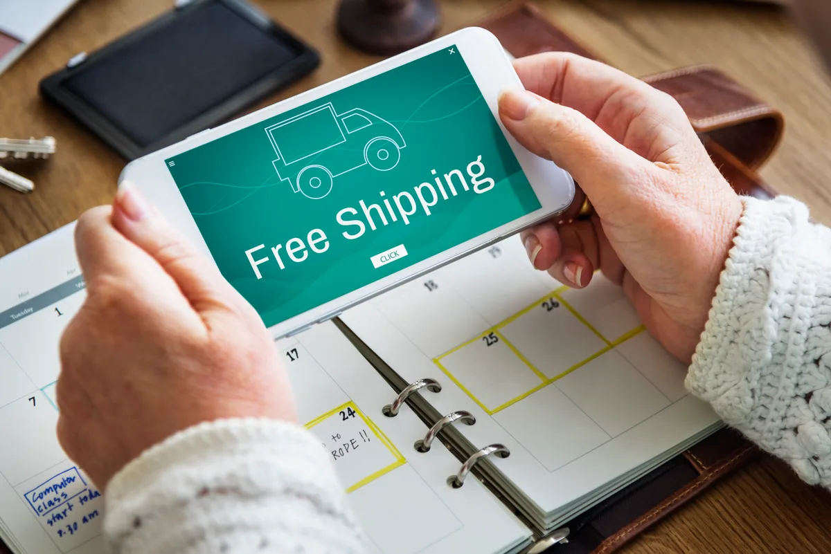 Poza cu doua maini daca tin un telefon in pozitie orizontala deasupra unui caiet iar pe ecranul telefonului scrie Free Shipping