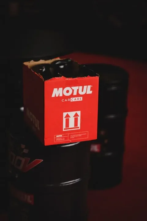 Poza cu o cutie rosie in care sunt puse produse Motul care sta pe doua butoaie de ulei Motul