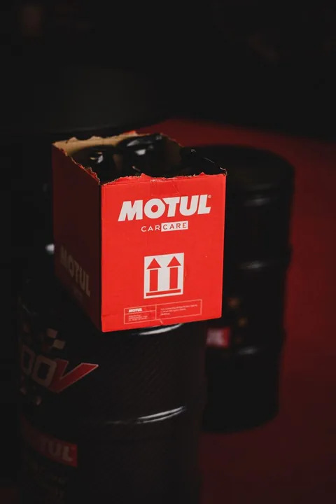 Poza cu o cutie rosie in care sunt puse produse Motul care sta pe doua butoaie de ulei Motul