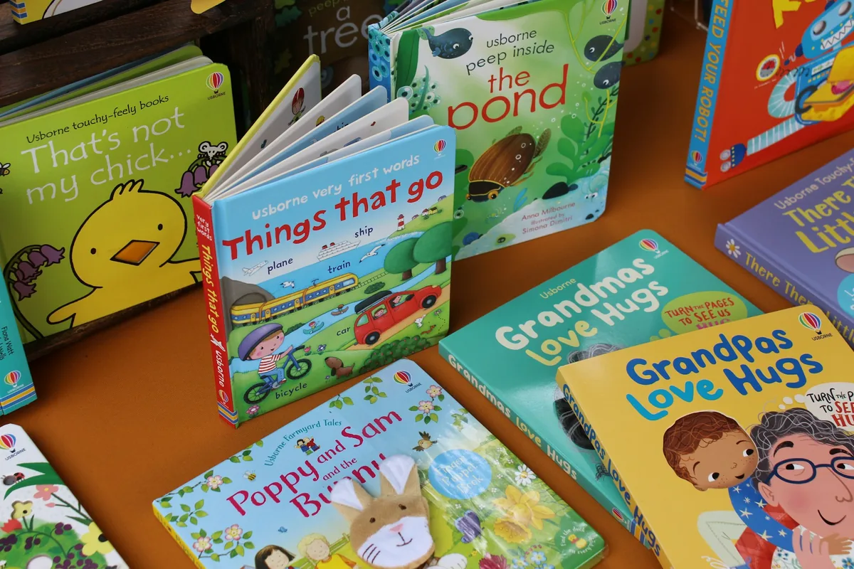 Poza cu mai multe carti destinate pentru copiii mici, model usborne asejate frumos cateva in picioare cateva culcate pe o masa de culoare maro