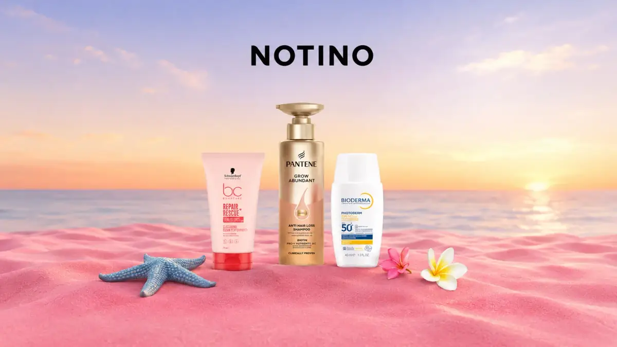 Produse cosmetice Notino pe nisip roz, cu flori tropicale și fundal de plajă la apus.
