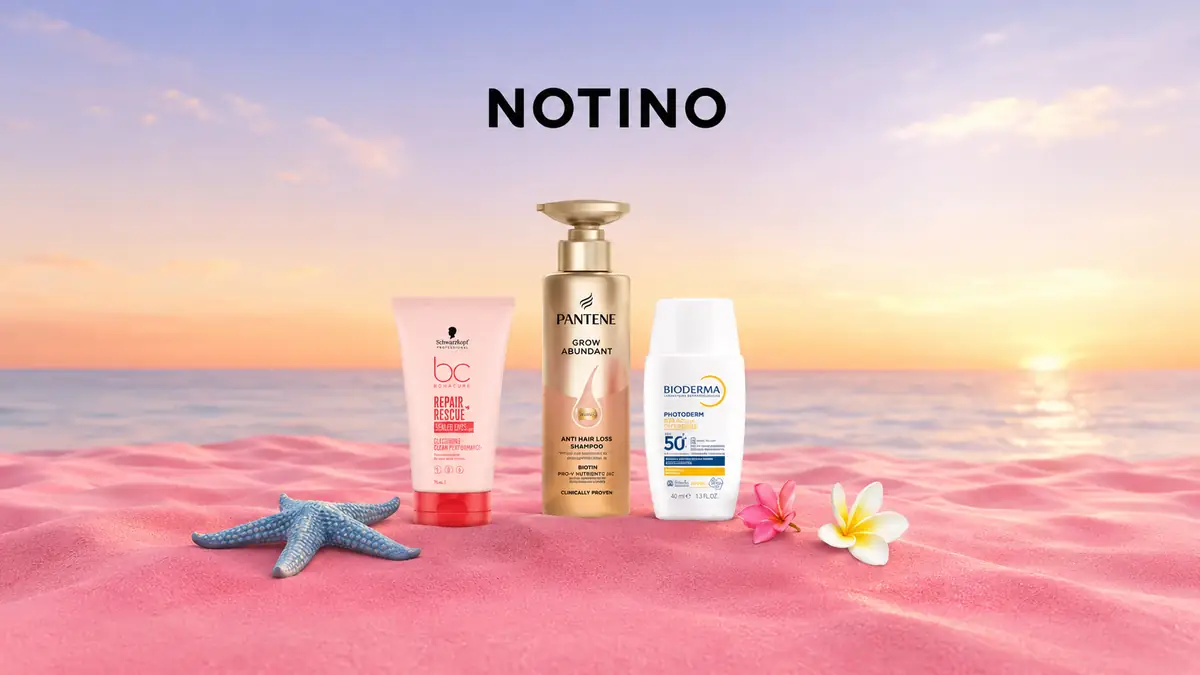 Produse cosmetice Notino pe nisip roz, cu flori tropicale și fundal de plajă la apus.
